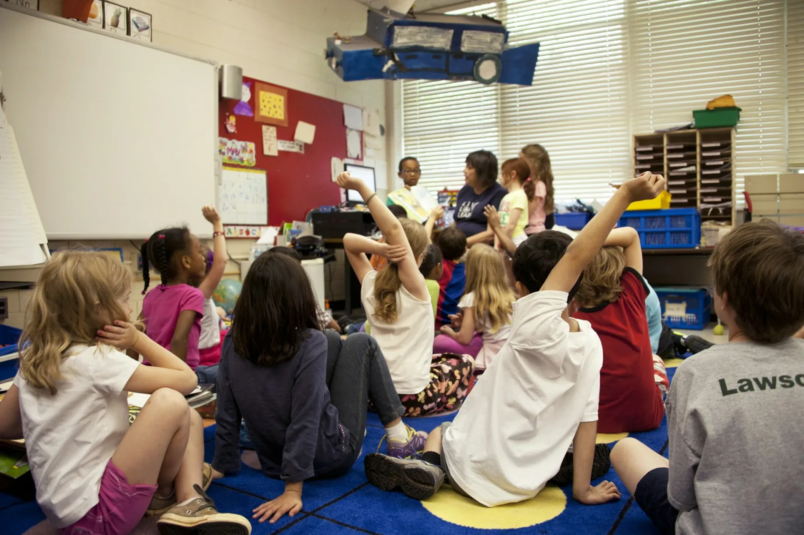 Elementary students gathered on a classroom rug during reading time, hands raised to answer