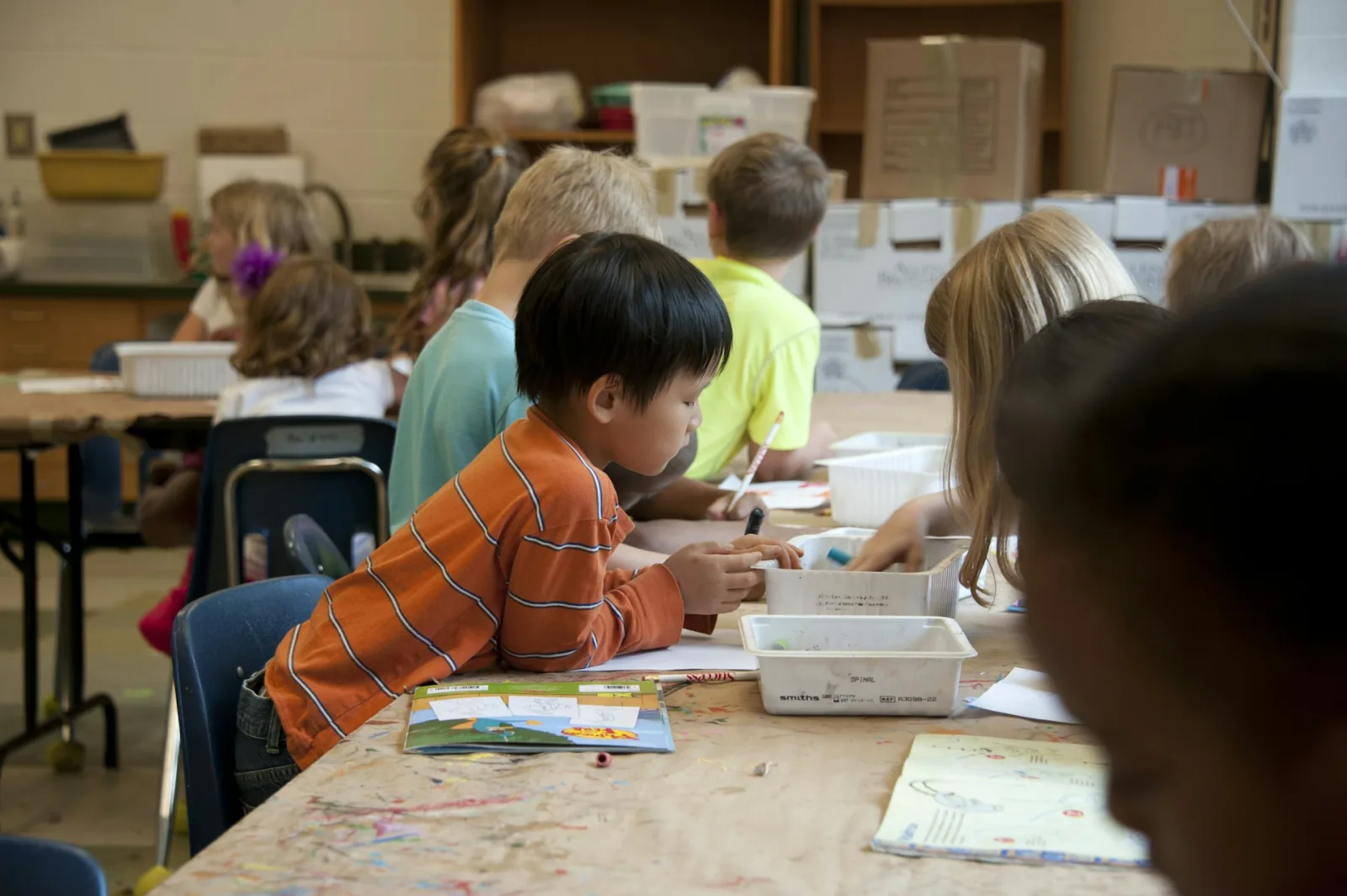 A young student working at a busy art table alongside his classmates